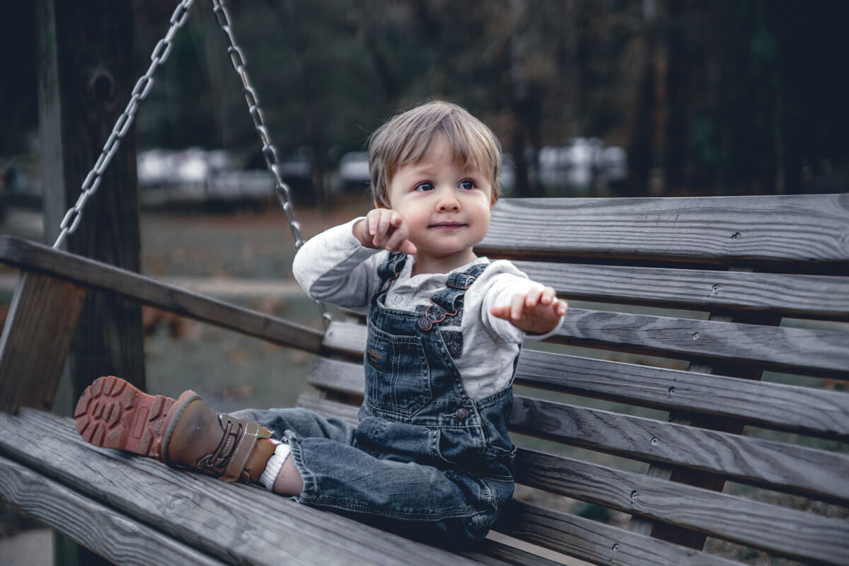 A toddler sitting on a swing holding out his hands to be picked up, everything but the toddler has had its colors deepened