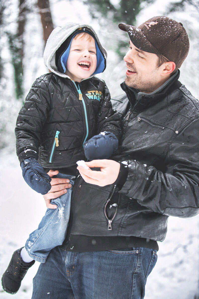 father holding 3 year old son and both laughing with a snowy background