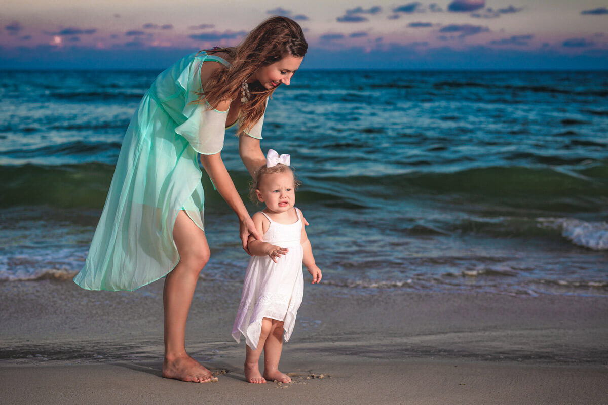 Mom helping toddler stand up on a sandy beach